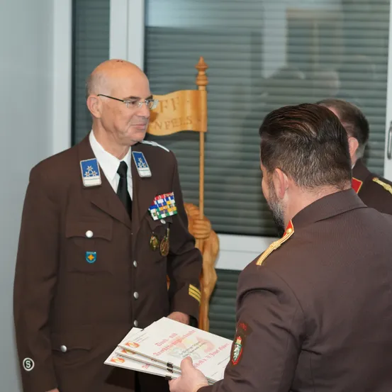 Ein uniformierter Offizier, der eine Brille trägt und eine Medaille trägt, gibt einem anderen Offizier ein Dokument, den er ansieht. Dahinter steht ein Holzpfahl mit einer Fahne.