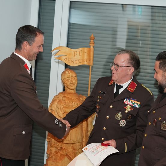 Zwei Männer in Militäruniformen schütteln sich die Hände. Einer hält ein Zertifikat, und eine Statue steht hinter ihnen.