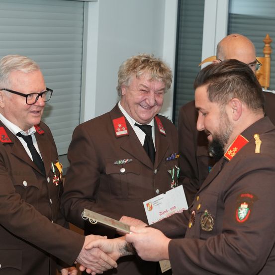 Drei Männer in Militäruniformen, einer präsentiert eine Plakette, ein anderer hält ein Zertifikat und der dritte lächelt, schüttelt Hände in einem Raum.