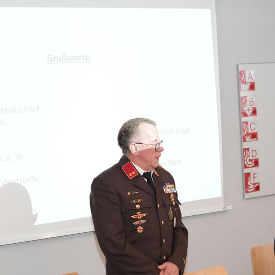 Ein Mann in Militäruniform steht vor einer Projektionsleinwand, wahrscheinlich in einem Klassenzimmer oder Konferenzraum. Er trägt eine Brille und eine Krawatte. Hinter ihm zeigt eine Tafel 'GruBworte' und der Schatten einer Person fällt auf die Wand.