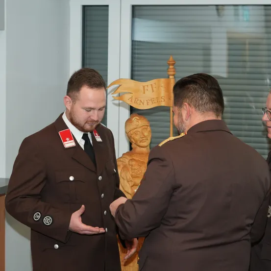 Zwei Männer in braunen Uniformen, einer mit einem roten Abzeichen, unterhalten sich in einem Raum mit einer hölzernen Statue im Hintergrund.
