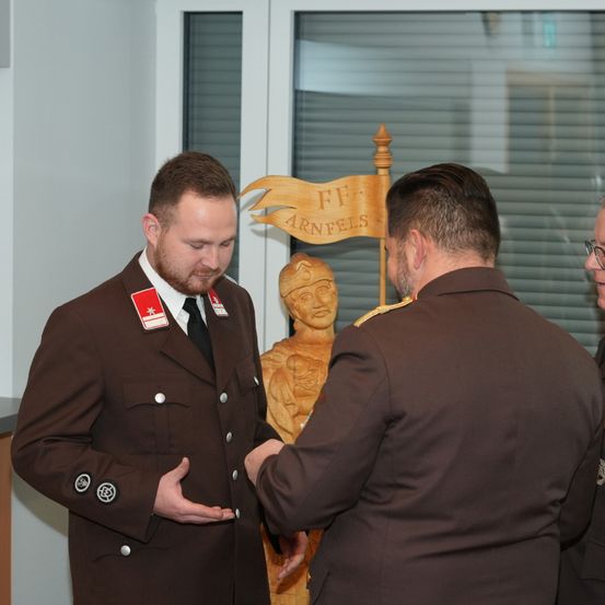 Zwei Männer in braunen Uniformen, einer mit einem roten Abzeichen, unterhalten sich in einem Raum mit einer hölzernen Statue im Hintergrund.