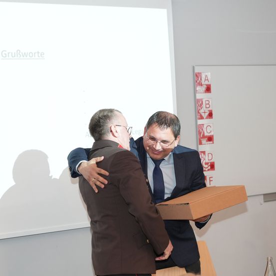 Zwei Männer stehen und umarmen sich, während einer eine Box in einem Raum mit einer Projektionsleinwand hält.