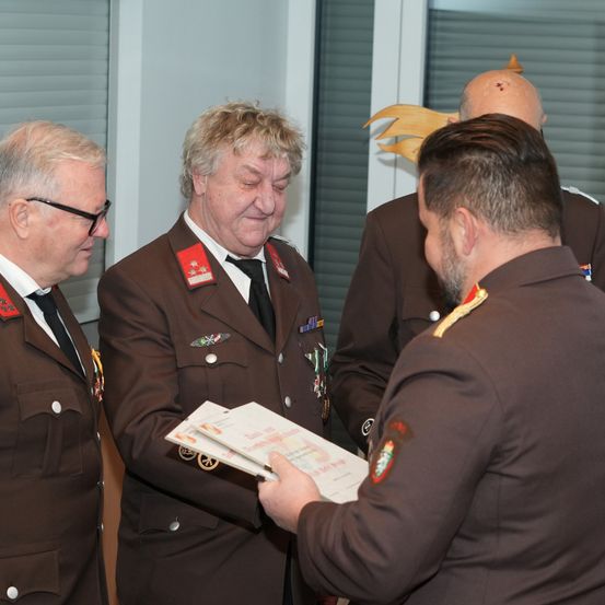Vier Männer in braunen Uniformen stehen drinnen. Ein Mann präsentiert einem anderen Mann ein Dokument. Die anderen beiden Männer beobachten.