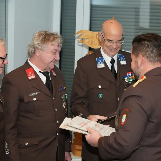 Vier Männer in Militäruniformen stehen und unterhalten sich. Ein Mann hält eine Urkunde und scheint sie einem anderen zu präsentieren. Hinter ihnen ist ein holznerner Pokal und ein Fenster mit Jalousien zu sehen.