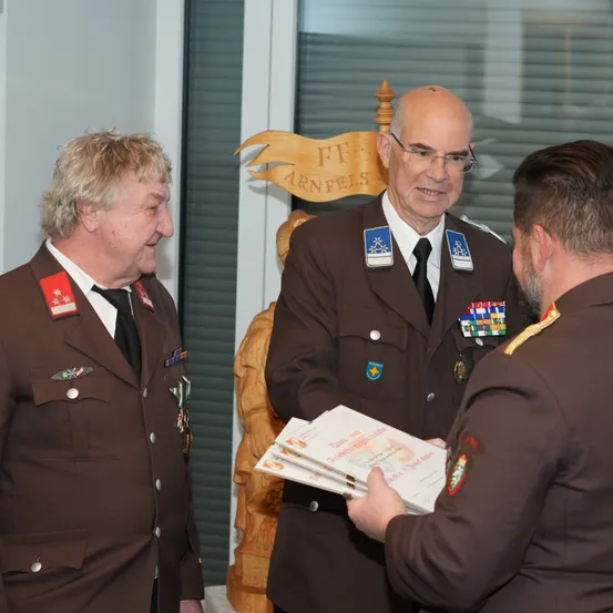 Drei Männer in braunen Uniformen stehen zusammen. Einer präsentiert einem anderen ein Dokument. Der Mann auf der rechten Seite trägt eine Brille und hat einen Schnurrbart. Hinter ihnen steht eine Holzskulptur.