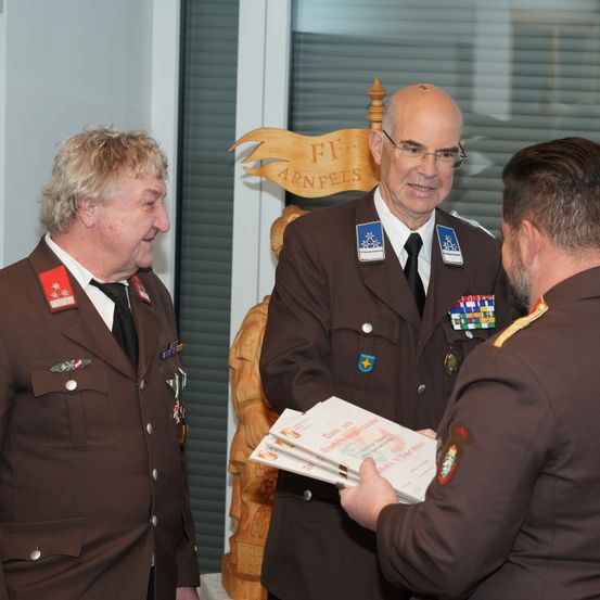 Drei Männer in braunen Uniformen stehen zusammen. Einer präsentiert einem anderen ein Dokument. Der Mann auf der rechten Seite trägt eine Brille und hat einen Schnurrbart. Hinter ihnen steht eine Holzskulptur.