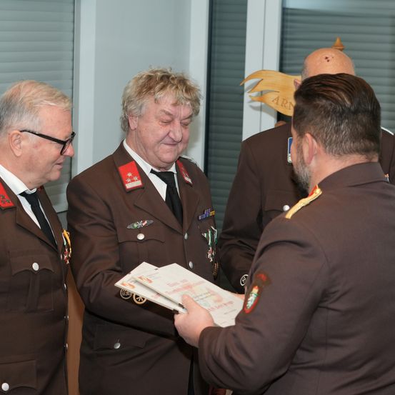 Vier Männer in Militäruniformen, einer hält ein Dokument, stehen in einem Raum mit Jalousien und einem Holzschild.