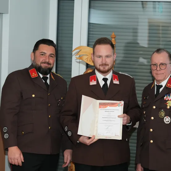 Drei Männer in Militäruniformen stehen nebeneinander, wobei der mittlere Mann ein Zertifikat in den Händen hält. Sie alle lächeln und scheinen für ein Foto zu posieren.