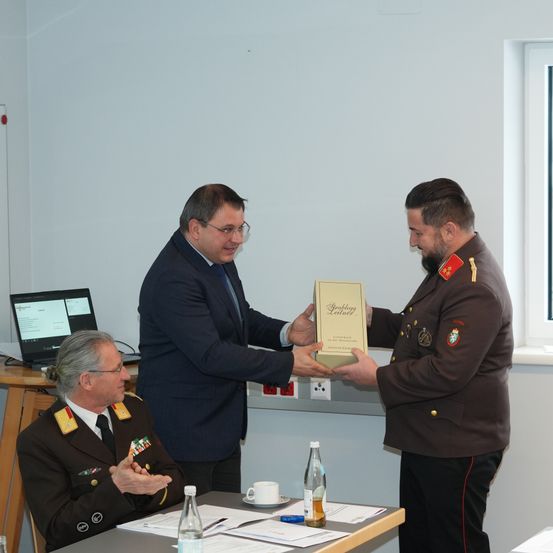 Drei Männer in Uniformen befinden sich in einem Besprechungsraum. Ein Mann in einem Anzug übergibt einem Militäroffizier ein gerahmtes Zertifikat. Papiere, Flaschen und ein Laptop sind auf dem Tisch.