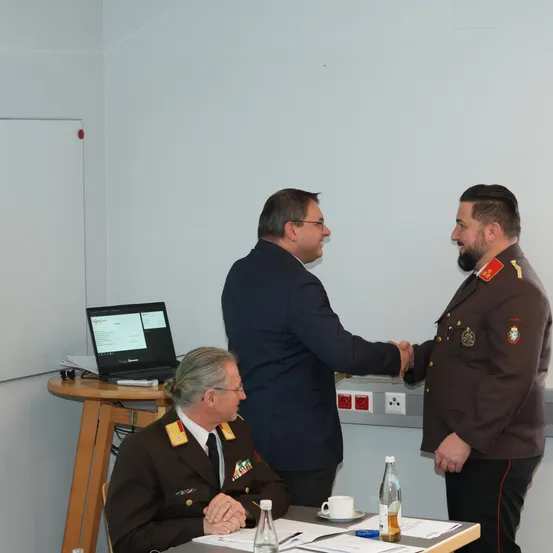 Drei Männer in Militäruniformen stehen in einem Raum, einer von ihnen schüttelt einem anderen die Hand. Ein Laptop steht auf einem Tisch neben ihnen.