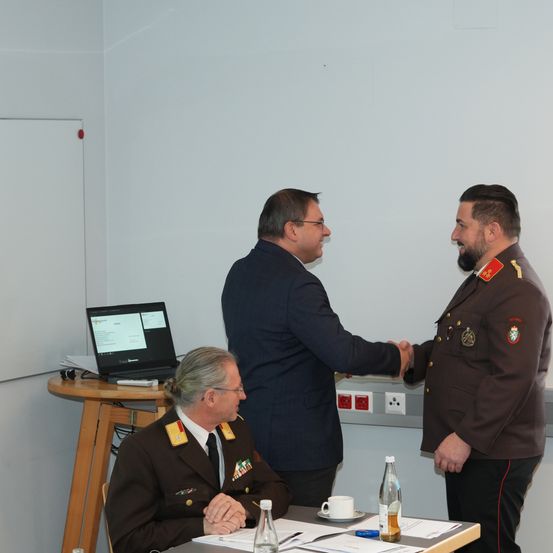 Drei Männer in Militäruniformen stehen in einem Raum, einer von ihnen schüttelt einem anderen die Hand. Ein Laptop steht auf einem Tisch neben ihnen.