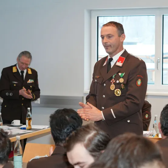 Ein Militäroffizier spricht zu einer Gruppe von Kadetten in einem Klassenzimmer, während er eine dekorierte Uniform trägt. Ein weiterer Offizier steht hinter ihm in der Nähe eines Tisches mit Flaschen und einem Becher.