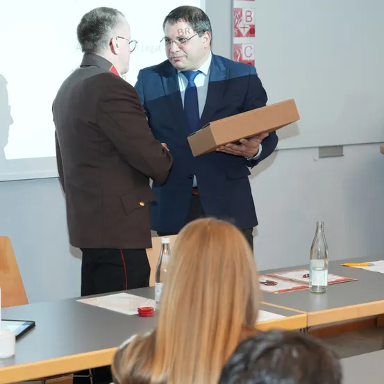 Zwei Männer stehen und unterhalten sich in einem Konferenzraum. Ein Mann hält eine Kartonbox, während andere an Tischen im Hintergrund sitzen.
