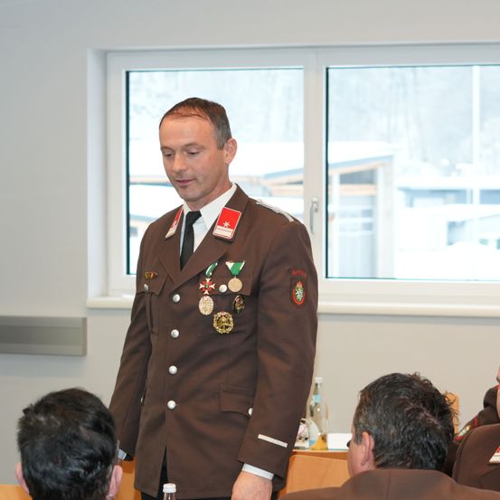 Ein Mann in einer braunen Uniform mit mehreren Medaillen steht vor einem Klassenzimmer. Der Raum ist gut beleuchtet mit Blick auf eine verschneite Landschaft draußen.