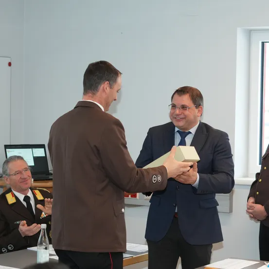 Ein Mann in einem braunen Anzug übergibt eine Schachtel an einen anderen Mann in einem blauen Anzug, beide lächeln. Ein dritter Mann in Uniform beobachtet, und ein Laptop steht auf einem Schreibtisch hinter ihnen.
