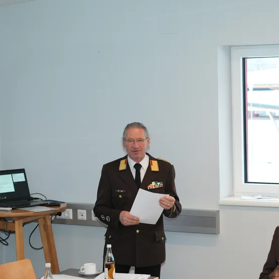 Ein Mann in Uniform steht vor einem Tisch mit einem Laptop und spricht zu einem Publikum. Hinter ihm befindet sich ein Fenster mit Blick nach draußen.