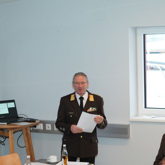 Ein Mann in Uniform steht vor einem Tisch mit einem Laptop und spricht zu einem Publikum. Hinter ihm befindet sich ein Fenster mit Blick nach draußen.