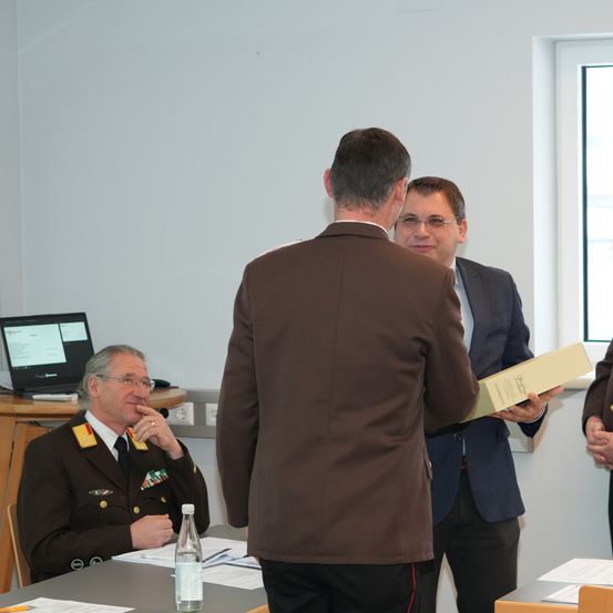 Drei Männer in formeller Kleidung, wahrscheinlich in einem Besprechungsraum. Einer steht und hält eine Mappe, ein anderer sitzt und lächelt, während der dritte steht und eine Brille trägt.