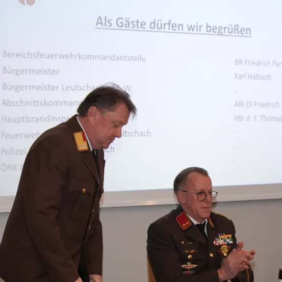 Zwei Männer in Militäruniformen stehen vor einem Bildschirm mit Text. Der Mann links sieht ernst aus, während der Mann rechts lächelt.