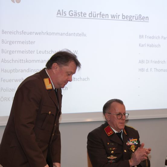 Zwei Männer in Militäruniformen stehen vor einem Bildschirm mit Text. Der Mann links sieht ernst aus, während der Mann rechts lächelt.