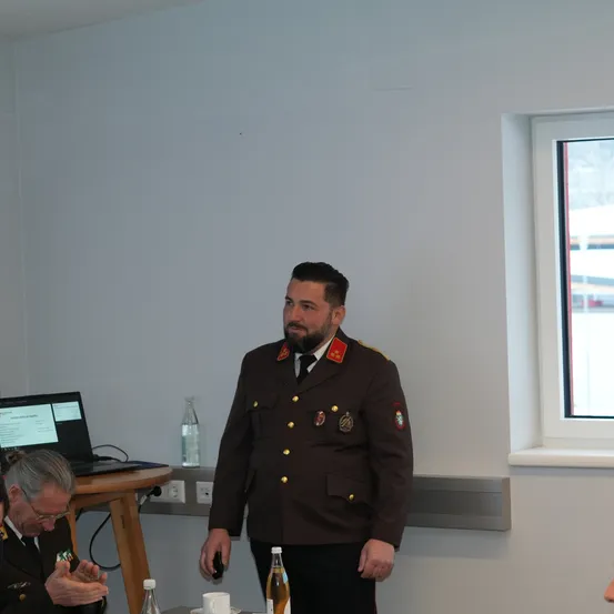 Ein Mann in Uniform steht in einem Raum mit Laptop, zwei Flaschen und Tassen auf einem Tisch. Ein älterer Mann in Uniform sitzt vor ihm. Ein Fenster rechts zeigt eine verschneite Szene draußen.