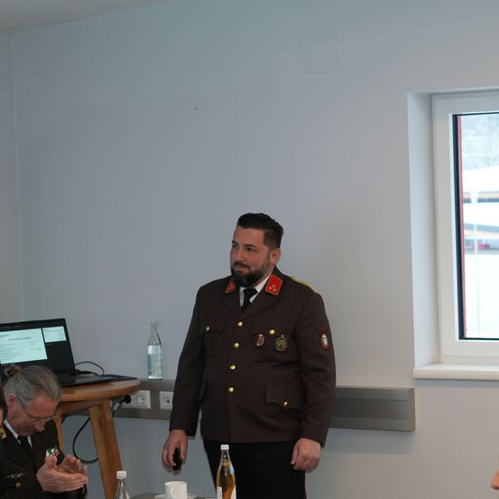 Ein Mann in Uniform steht in einem Raum mit Laptop, zwei Flaschen und Tassen auf einem Tisch. Ein älterer Mann in Uniform sitzt vor ihm. Ein Fenster rechts zeigt eine verschneite Szene draußen.
