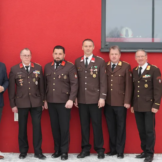 Fünf Männer in Militäruniformen stehen vor einer roten Wand. Sie posieren für ein Foto mit ernsten Gesichtern. Hinter ihnen ist ein Glasfenster mit der Reflexion eines Helms.