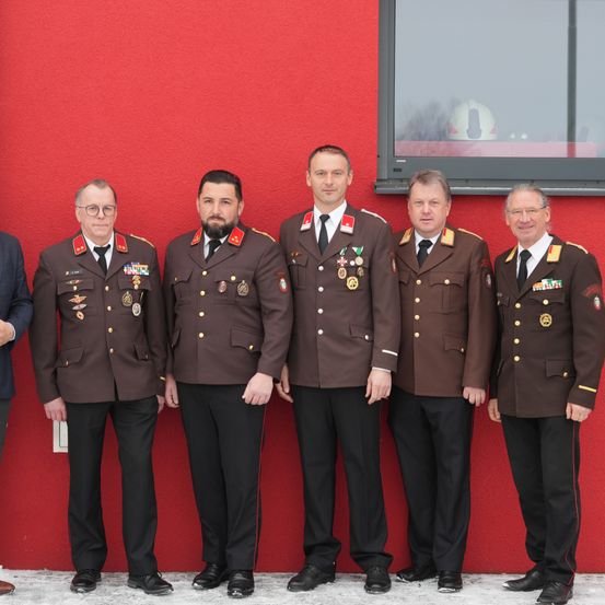 Fünf Männer in Militäruniformen stehen vor einer roten Wand. Sie posieren für ein Foto mit ernsten Gesichtern. Hinter ihnen ist ein Glasfenster mit der Reflexion eines Helms.