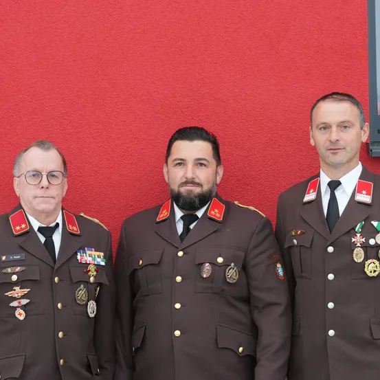 Drei Männer in Militäruniformen mit Medaillen stehen vor einer roten Wand. Der Mann auf der linken Seite trägt eine Brille. Der Mann in der Mitte hat einen Bart und einen Schnurrbart. Der Mann auf der rechten Seite hat einen roten Stern auf seiner Schulter.