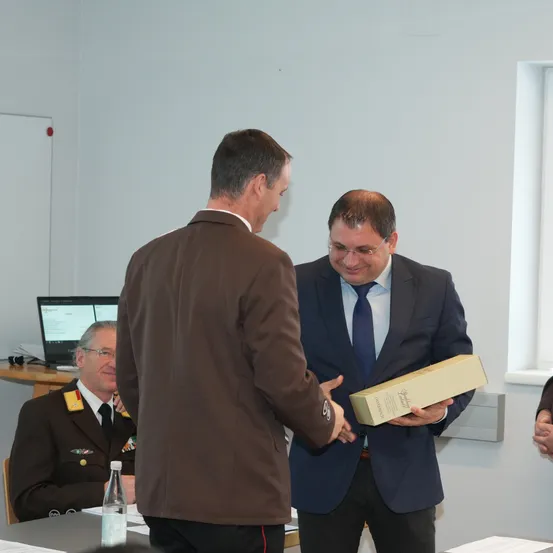 Zwei Männer stehen in einem Besprechungsraum und schütteln sich die Hände. Einer hält eine Box. Ein weiterer Mann in Uniform sitzt und lächelt. Es gibt einen Tisch mit einer Flasche und Papieren.