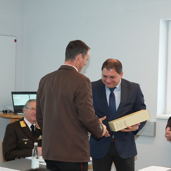 Zwei Männer stehen in einem Besprechungsraum und schütteln sich die Hände. Einer hält eine Box. Ein weiterer Mann in Uniform sitzt und lächelt. Es gibt einen Tisch mit einer Flasche und Papieren.