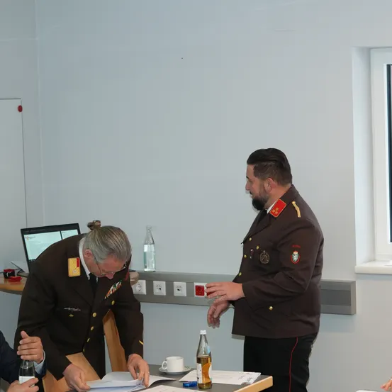 Zwei Männer in Militäruniformen unterhalten sich in der Nähe eines Schreibtisches, auf dem ein Laptop und Flaschen stehen. Der Raum hat weiße Wände und ein offenes Fenster.