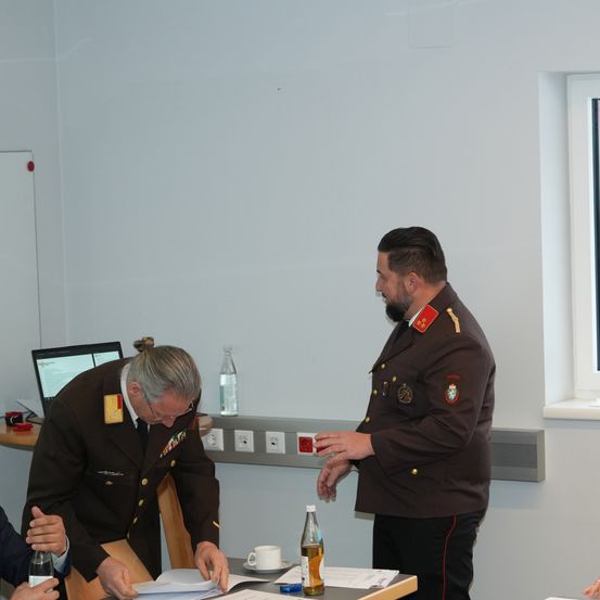 Zwei Männer in Militäruniformen unterhalten sich in der Nähe eines Schreibtisches, auf dem ein Laptop und Flaschen stehen. Der Raum hat weiße Wände und ein offenes Fenster.