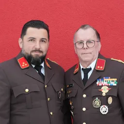 Zwei Männer in Militäruniformen stehen vor einer roten Wand. Der Mann auf der linken Seite hat einen Bart und trägt einen roten Stern auf seiner Schulter. Der Mann auf der rechten Seite trägt eine Brille und hat Medaillen auf seiner Brust.