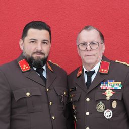 Zwei Männer in Militäruniformen stehen vor einer roten Wand. Der Mann auf der linken Seite hat einen Bart und trägt einen roten Stern auf seiner Schulter. Der Mann auf der rechten Seite trägt eine Brille und hat Medaillen auf seiner Brust.