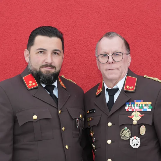 Zwei Männer in Militäruniformen stehen vor einer roten Wand. Der Mann auf der linken Seite hat einen Bart und trägt einen roten Stern auf seiner Schulter. Der Mann auf der rechten Seite trägt eine Brille und hat Medaillen auf seiner Brust.