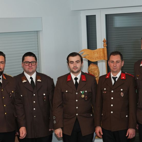 Fünf Männer in braunen Uniformen, möglicherweise Militäroffiziere, stehen in einem Raum mit einer weißen Wand und einem Fenster. Der Mann in der Mitte hat Medaillen auf der Brust.