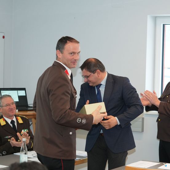 Ein Mann in einer braunen Militäruniform überreicht einem Mann in einem blauen Anzug eine Schachtel. Hinter ihnen klatschen zwei weitere Personen. Ein Laptop und eine Flasche stehen auf einem Tisch.