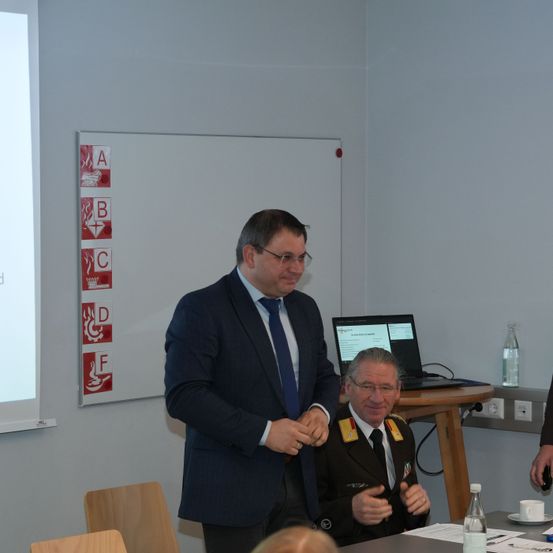 Zwei Männer in formeller Kleidung sitzen vor einem Schreibtisch und unterhalten sich. Hinter ihnen befindet sich eine Tafel mit Sicherheitssymbolen. Ein Laptop steht auf dem Schreibtisch, und es gibt eine Tasse und eine Flasche. Ein weiterer Mann steht in der Nähe und hört ihnen möglicherweise zu.