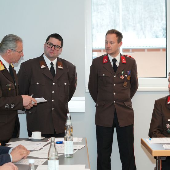 Vier Männer in Militäruniformen stehen in einem Raum. Drei von ihnen stehen sich gegenüber, während der vierte hinter einem Tisch steht. Papiere, Stifte und Wasserflaschen liegen auf dem Tisch.