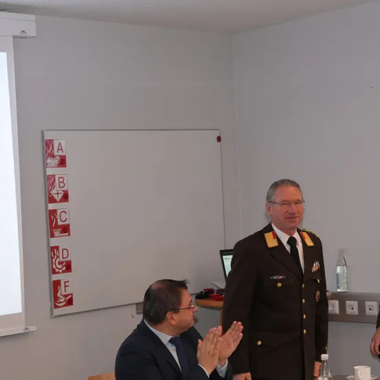 Zwei Männer sind in einem Konferenzraum. Einer klatscht, während der andere steht und lächelt. Hinter ihnen befindet sich eine Tafel mit Sicherheitszeichen.
