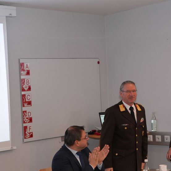 Zwei Männer sind in einem Konferenzraum. Einer klatscht, während der andere steht und lächelt. Hinter ihnen befindet sich eine Tafel mit Sicherheitszeichen.