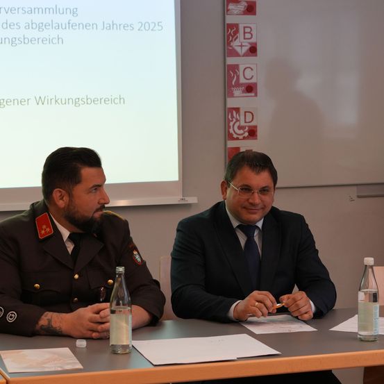 Zwei Männer sitzen an einem Tisch in einem, was wie ein Besprechungsraum aussieht. Einer ist in Uniform, und der andere ist in einem Anzug. Papiere und Flaschen sind auf dem Tisch. Hinter ihnen ist eine Projektionsleinwand, die Text auf Deutsch anzeigt.