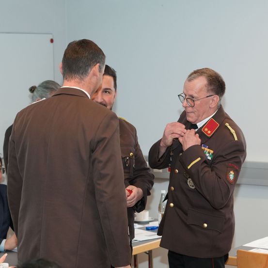 Ein Mann in Uniform befestigt seine Krawatte und schaut zwei Männer vor sich an. Ein anderer Mann lächelt und schaut auf den Mann, der seine Krawatte festmacht. Dahinter steht ein Tisch mit einer Flasche, einem Becher und einem Teller. Eine Frau steht hinter ihnen.