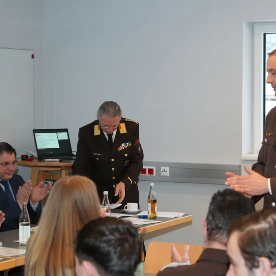 Ein Mann in Uniform steht an einem Tisch mit einem Laptop und spricht zu einem Publikum. Das Publikum hört aufmerksam zu, einige klatschen. Ein anderer Mann in Uniform steht neben ihm. Der Raum ist einfach eingerichtet mit einer Tafel, Steckdosen und einem Fenster.