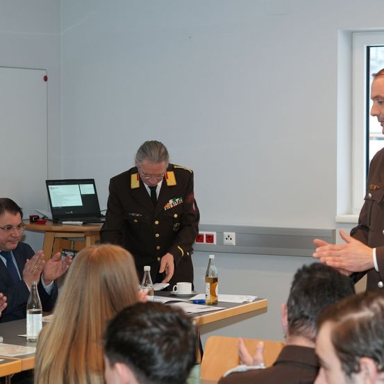 Ein Mann in Uniform steht an einem Tisch mit einem Laptop und spricht zu einem Publikum. Das Publikum hört aufmerksam zu, einige klatschen. Ein anderer Mann in Uniform steht neben ihm. Der Raum ist einfach eingerichtet mit einer Tafel, Steckdosen und einem Fenster.