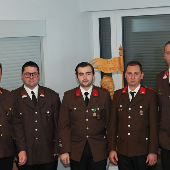 Fünf Männer in Militäruniformen posieren für ein Foto in einem Raum. Hinter ihnen eine Fahne mit Text und eine geschnitzte Holzfigur.