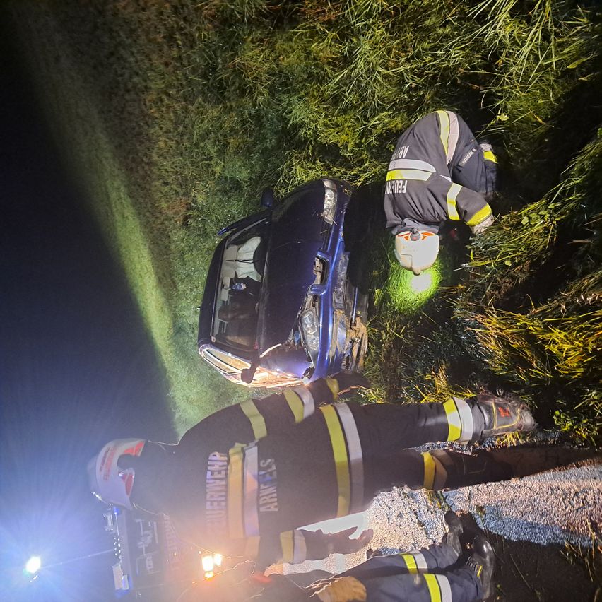 Feuerwehrleute in Schutzkleidung retten jemanden aus einem zerstörten blauen Auto an einem grasbewachsenen Straßenrand bei Nacht.
