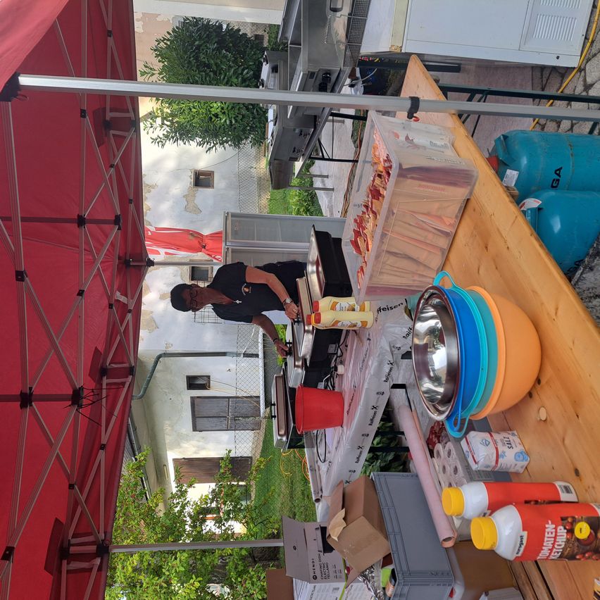 Eine Person bereitet Essen unter einem roten Baldachin zu. Ein Schirm, Propangasflaschen und eine Schüssel stehen auf dem Tisch. Ein weißes Gebäude ist im Hintergrund.